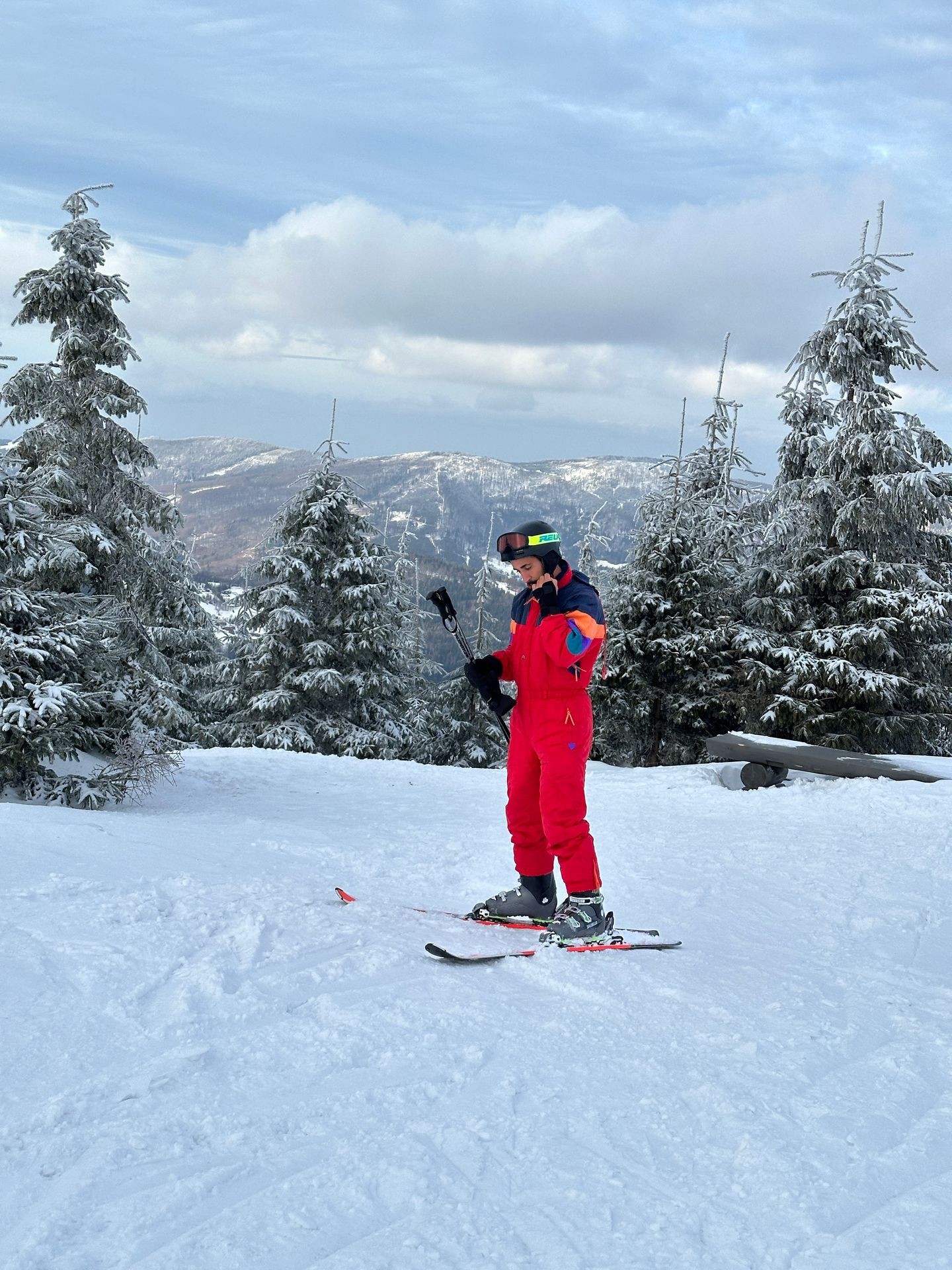 Person in red ski suit standing on snowy mountain surrounded by snow-covered trees, holding ski poles.