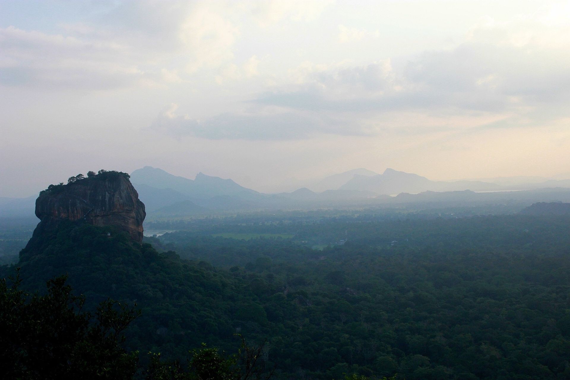 Scenic view of a rocky hill surrounded by lush greenery and distant mountains under a cloudy sky.