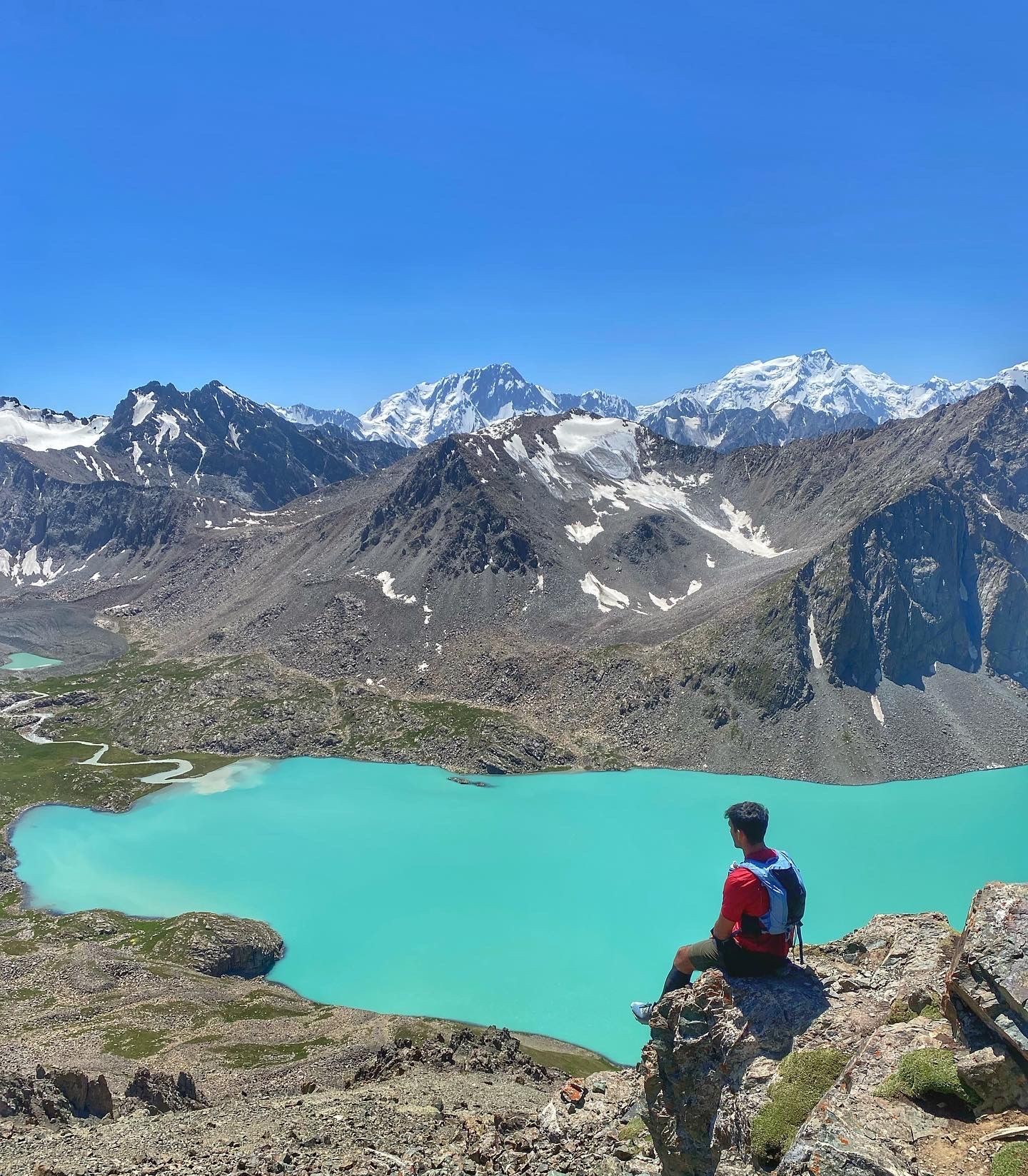 Person sitting on a rock overlooking a turquoise lake surrounded by snowy mountains under a clear blue sky.